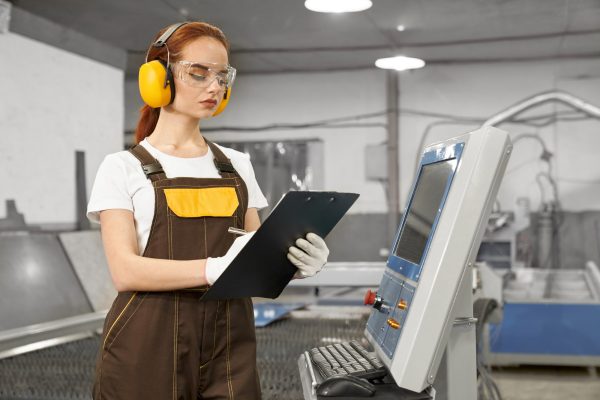 Serious, concentrated female engineer holding folder, operating computerized plasma laser machine. Beautiful woman wearing in white t shirt, gloves, coveralls, protective ear muffs and glasses.